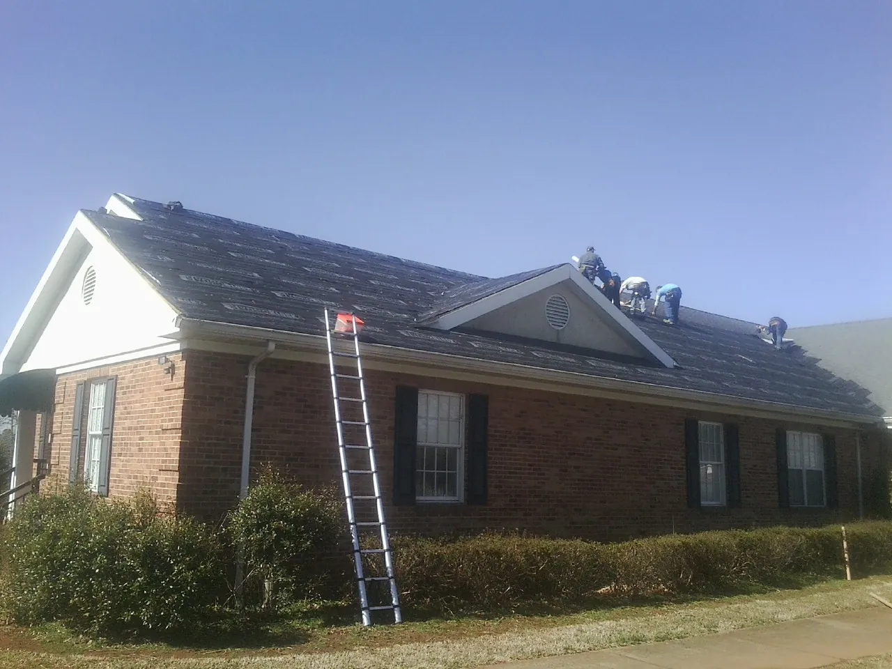 Crew tearing shingles off a brick ranch roof with a ladder set — Emergency Roof Repair work by our crew in Quinby