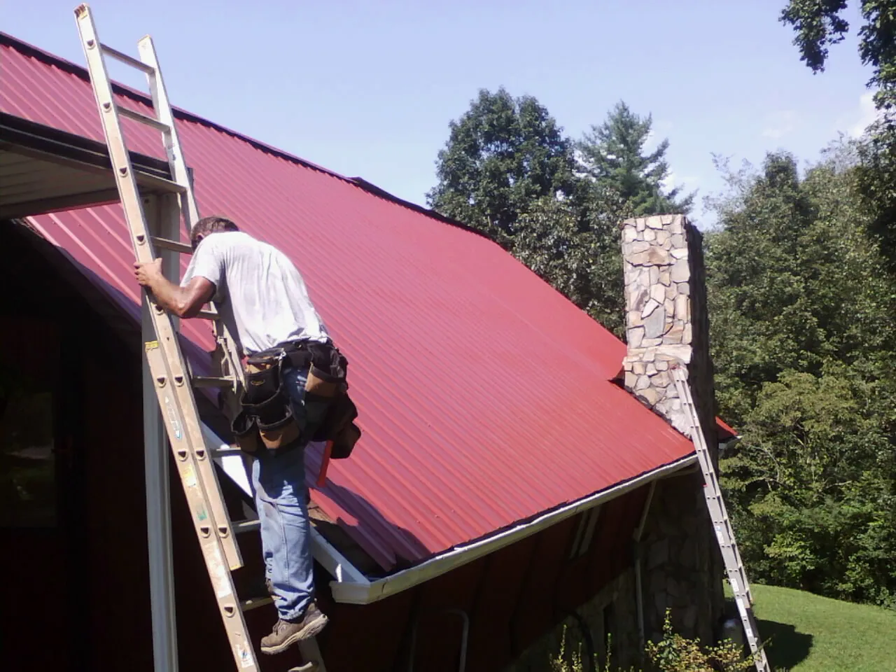 Roofer on a ladder beside a red metal roof and stone chimney — Soffit & Fascia Repair by Roofing Brothers in Quinby